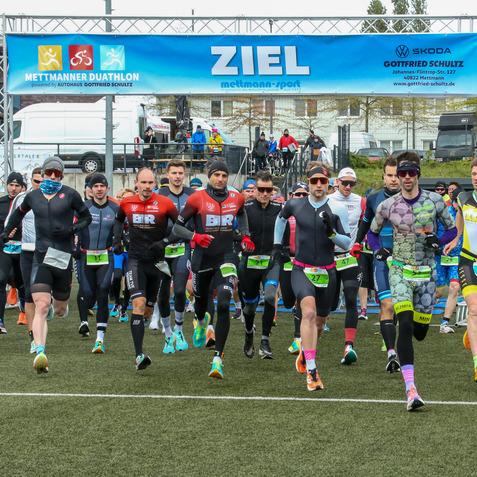 Duathlon 2024 Start des Mettmanner Duathlons: Läufer in Sportkleidung laufen auf einem Rasenfeld unter einem Zielbanner.