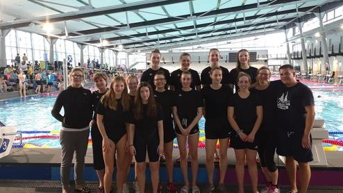 Gruppenfoto von Schwimmerinnen in einheitlichen schwarzen Badeanzügen vor einem Schwimmbecken in einem modernen Hallenbad.