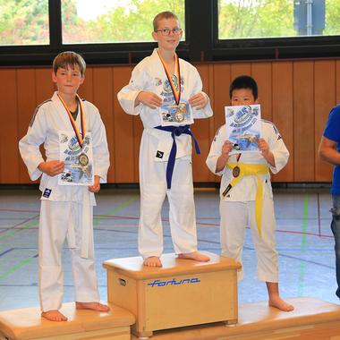 Drei Kinder in Karateanzügen stehen auf dem Siegertreppchen, halten Medaillen und Urkunden, während ein Erwachsener zusieht.