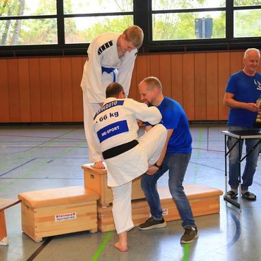 Judo-Kämpfer beim Training, unterstützt von einem Trainer, während Trophäen am Tisch aufgestellt sind.