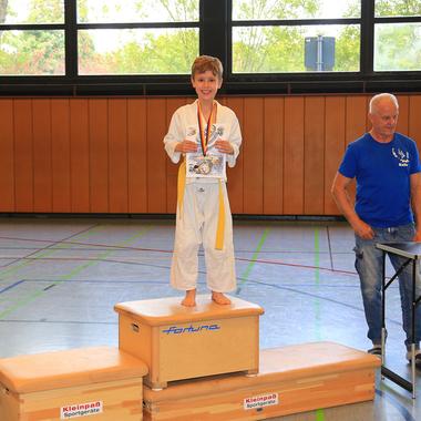 Junge in Karateanzug steht auf dem Siegerpodest, hält Medaille und Urkunde, während Trainer neben Pokalen lächelt.