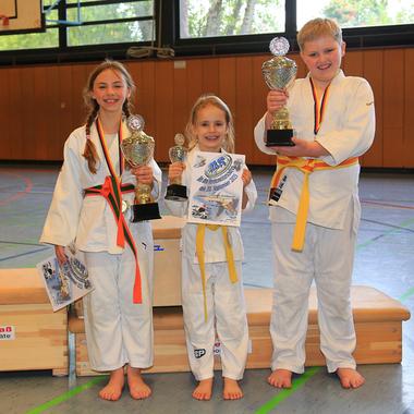 Drei Kinder in Karateanzügen halten Pokale und eine Urkunde auf einer Sportfläche.