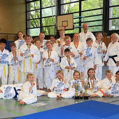 Gruppe junger Judoka in weißen Anzügen mit Medaillen und Urkunden, versammelt in einer Sporthalle.
