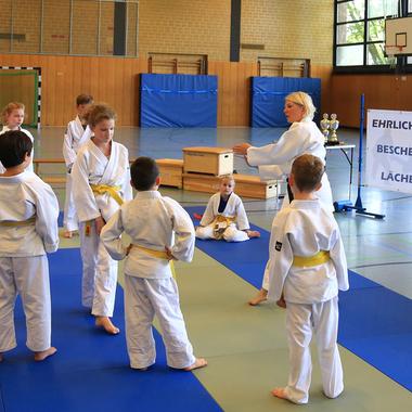 Gruppe von Kindern in Judoanzügen hört einer Trainerin in einem Judo-Trainingsraum zu, mit Pokalen im Hintergrund.