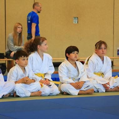 Sechs Kinder im Judogi sitzen auf Matten, aufmerksam einem Trainer in blauem Shirt zuhörend.
