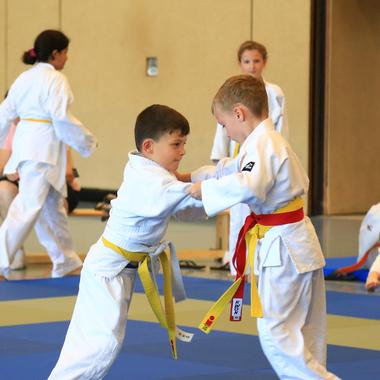 Zwei Kinder im weißen Judoanzug üben einen greifenden Wurf auf einer blauen Judomatte, mit einem weiteren Kind im Hintergrund.