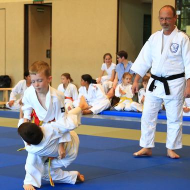 Zwei Kinder in Judoanzügen üben eine Technik auf einer blauen Judomatte, während ein Erwachsener zusieht.