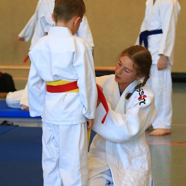 Trainerin bindet einem jungen Judoka den roten Gürtel an, während im Hintergrund weitere Kinder in Judokleidung stehen.