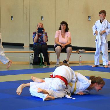 Zwei Judokas in weißen Anzügen üben auf einer blauen Matte, während Zuschauer im Hintergrund entspannt sitzen.