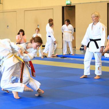 Zwei Kinder im Judojacket kämpfen auf einer blauen Matte, während Beobachter im Hintergrund zuschauen.