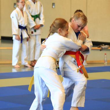 Zwei Kinder im Judo-Anzug kämpfen auf einer blauen Matte, während andere Kinder zuschauen.