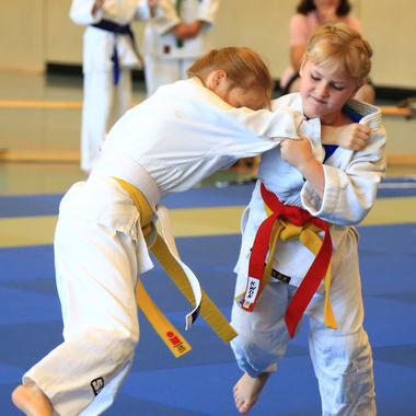 Zwei Kinder im Judogi kämpfen auf einer blauen Judomatte, während Zuschauer im Hintergrund aufgeregt zuschauen.