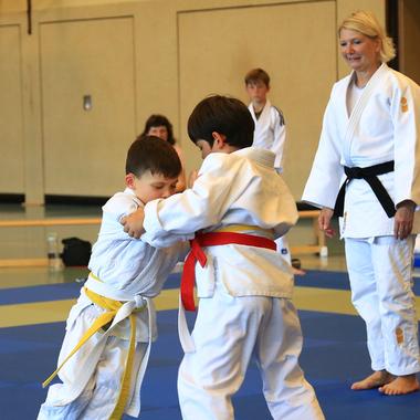 Zwei Kinder im Judoanzug kämpfen spielerisch auf einer blauen Judomatte, während ein Erwachsener zuschaut.