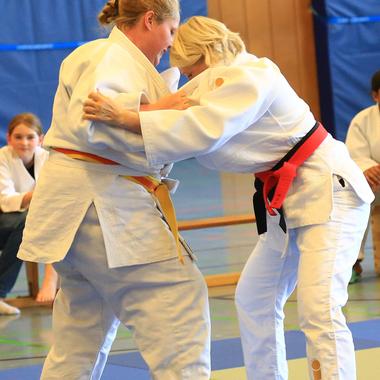 Zwei Frauen im Judo-Anzug üben einen Griff auf einer Judomatte, Zuschauer sitzen im Hintergrund.