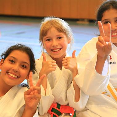 Drei Mädchen in Judoanzügen zeigen Daumen hoch und V-Zeichen in einer Sporthalle.