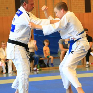 Zwei Judoka in Weiß und Schwarz kämpfen auf einer blauen Matte; im Hintergrund sitzen Zuschauer.
