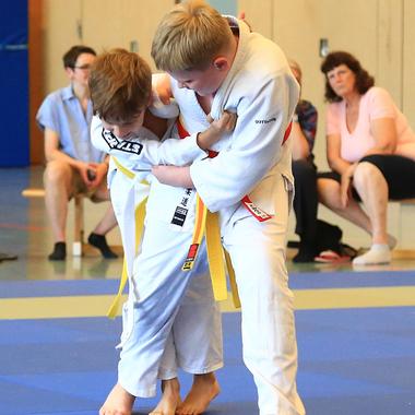 Zwei Kinder im Judokampf, beide in weißen Gi mit gelben Gürteln, auf einer blauen Matte in einer Sporthalle.