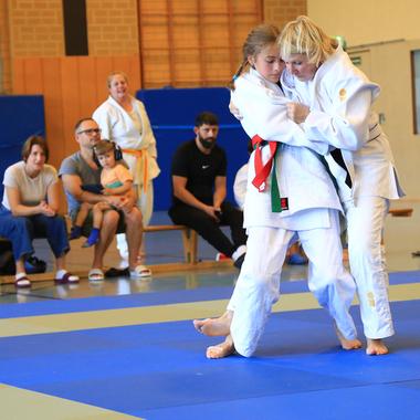 Zwei Judoka in Weiß und Schwarz kämpfen auf einer blauen Matte, während Zuschauer im Hintergrund zuschauen.