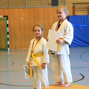 Zwei Mädchen in Judoanzügen stehen auf einem Siegertreppchen, eines hält eine Urkunde, das andere eine Medaille.