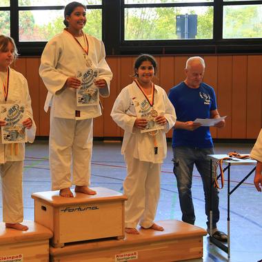 Drei Karateka stehen auf dem Siegertreppchen mit Medaillen und Urkunden, während ein Zuschauer applaudiert.