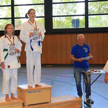 Zwei Sportlerinnen stehen auf dem Siegerpodest mit Medaillen und Urkunden in einer Turnhalle.