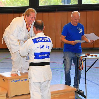Junge Frau steht auf einem Podest, erhält Medaille; zwei Männer applaudieren, Trophäen und Urkunden auf Tisch.