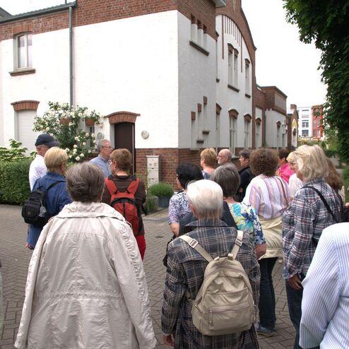 Gruppe von Menschen hört einem Guide vor einem Gebäude mit weißen Wänden und Blumen zu.