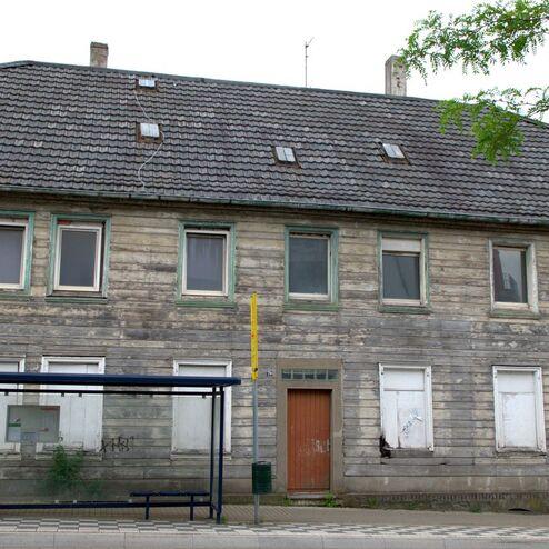Verfallenes Holzhaus mit grauem Dach und geschlossenen Fenstern, darunter eine Haltestelle mit Wartebank.