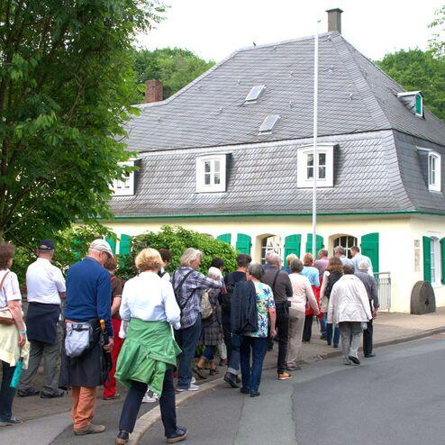 Gruppe von Menschen, die an einem Haus mit grünen Fensterläden entlanggeht, umgeben von Bäumen und Sträuchern.