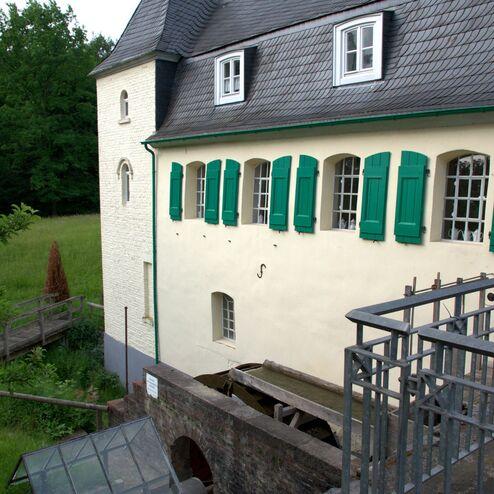Fassade eines historischen Gebäudes mit einem runden Turm, grünen Fensterläden und Sicht auf ein Wasserrad.