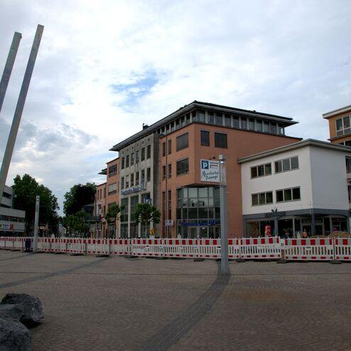 Stadtplatz mit Gebäuden, Baustellenabsperrungen und einer Skulptur im Vordergrund unter bewölktem Himmel.