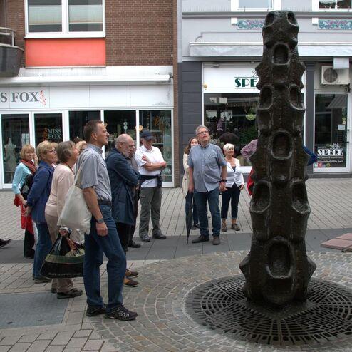 Gruppe von Menschen betrachtet eine hohe Skulptur auf einem Pflasterweg in einer Fußgängerzone.
