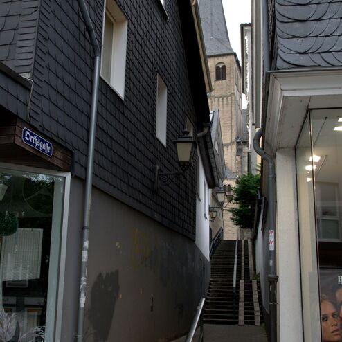 Enge Gasse mit Treppen zwischen alten Häusern und Blick auf den Kirchturm im Hintergrund.