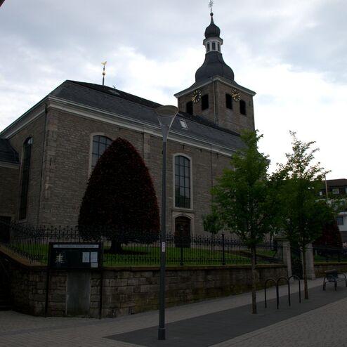 Historisches Gebäude mit Turm und großen Fenstern, umgeben von Bäumen und einer gepflasterten Straße.