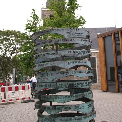 Skulptur mit geschwungenen Metallstreifen, die Jubiläen und Veranstaltungen in Mettmann anzeigt, umgeben von Bäumen.
