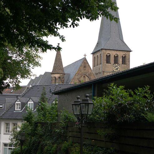 Blick auf eine Kirche mit spitzem Turm, umgeben von Bäumen und Wohnhäusern in einer ruhigen Straße.
