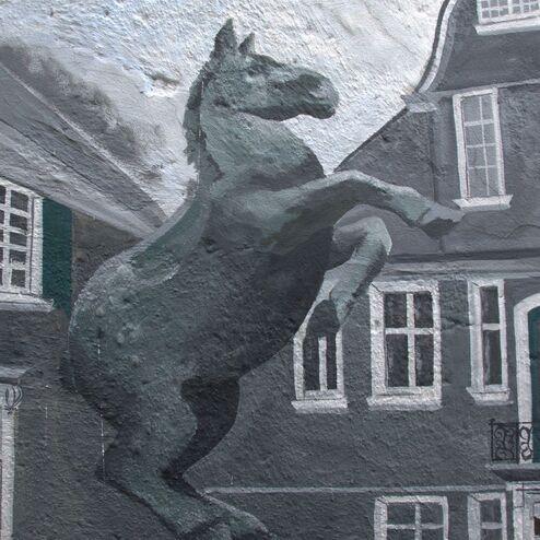 Wandmalerei eines aufrecht stehenden Pferdes vor einem grauen Gebäude mit Fenstern.
