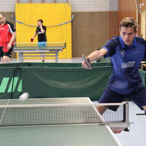 Junge spielt Tischtennis, während zwei weitere Spieler im Hintergrund auf einer grünen Tischtennisplatte aktiv sind.