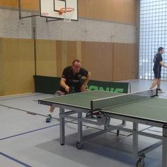 Ein Spieler beim Tischtennis steht an einem Tisch und bereitet sich auf einen Schlag vor, im Hintergrund weitere Spieler.