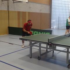 Tischtennisspieler in roter Kleidung schlägt den Ball auf einer Tischtennisplatte in einer Sporthalle.