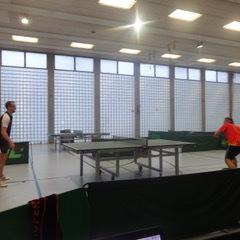 Zwei Tischtennisspieler in einem sportlichen Wettkampf an einem Tischtennistisch in einer hellen Halle.