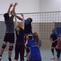 Zwei Spieler springen, um einen Volleyball über das Netz zu blocken, während weitere Spieler im Hintergrund beobachten.