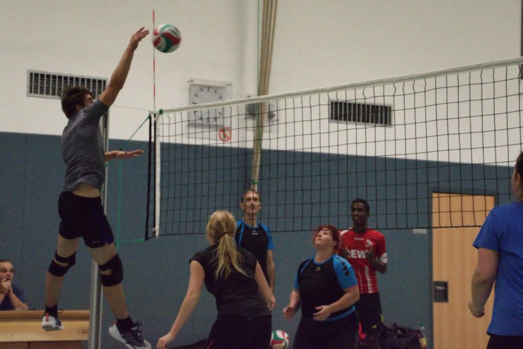 Spieler springt hoch, um einen Volleyball über das Netz zu schlagen, während andere Teammitglieder beobachten.