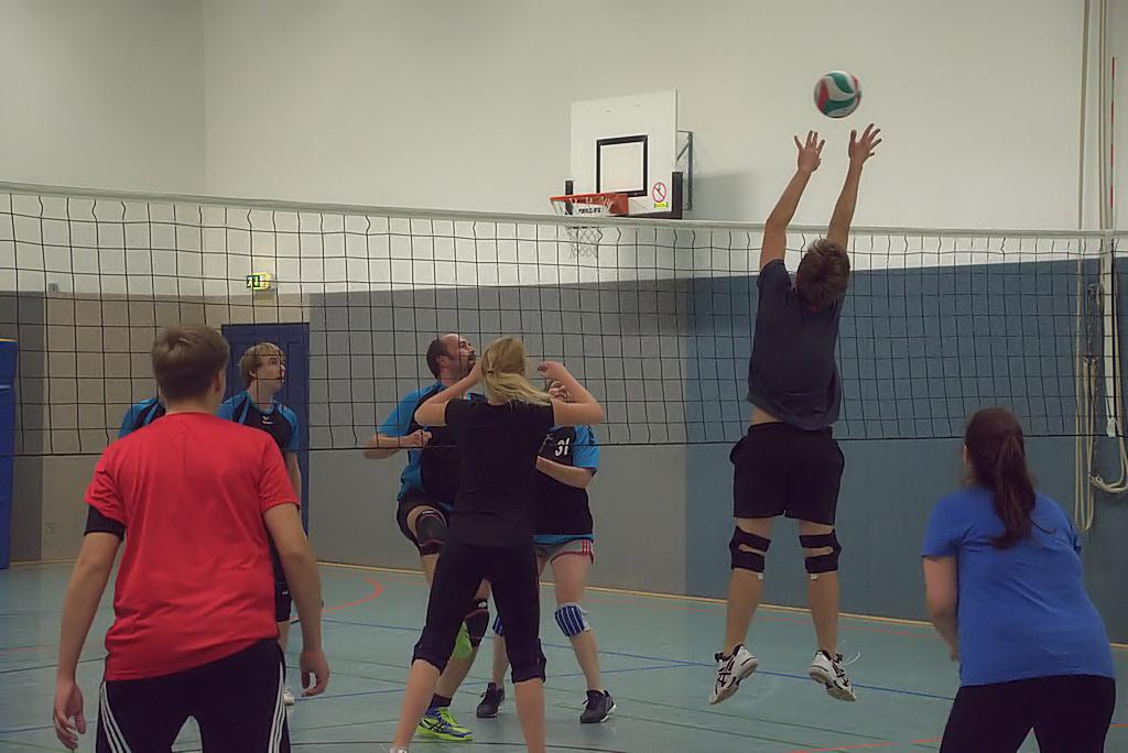 Spielszene im Volleyball: Ein Spieler springt hoch, um einen Ball über das Netz zu blocken, während andere zuschauen.