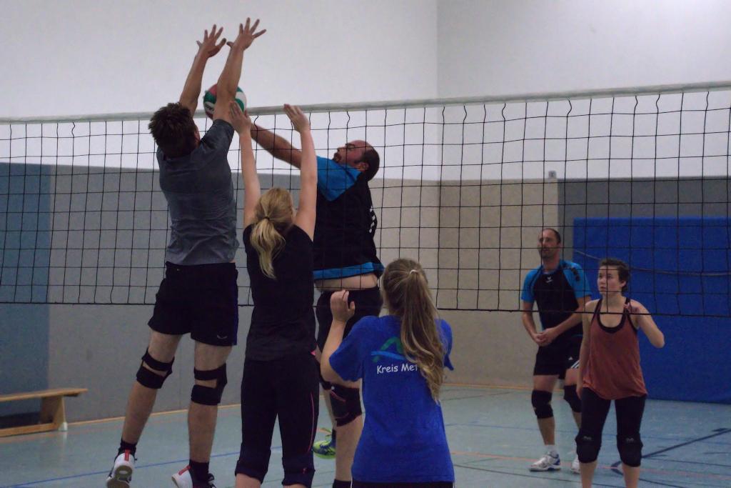 Zwei Spieler springen, um einen Volleyball über das Netz zu blocken, während weitere Spieler im Hintergrund beobachten.