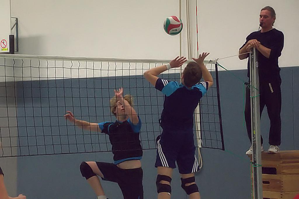 Zwei Volleyballspieler springen zum Ball über einem Netz, während ein Schiedsrichter auf einem Stuhl beobachtet.
