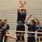 Zwei Volleyballspieler blocken am Netz, während ein dritter Spieler im Hintergrund den Ball anspielt.