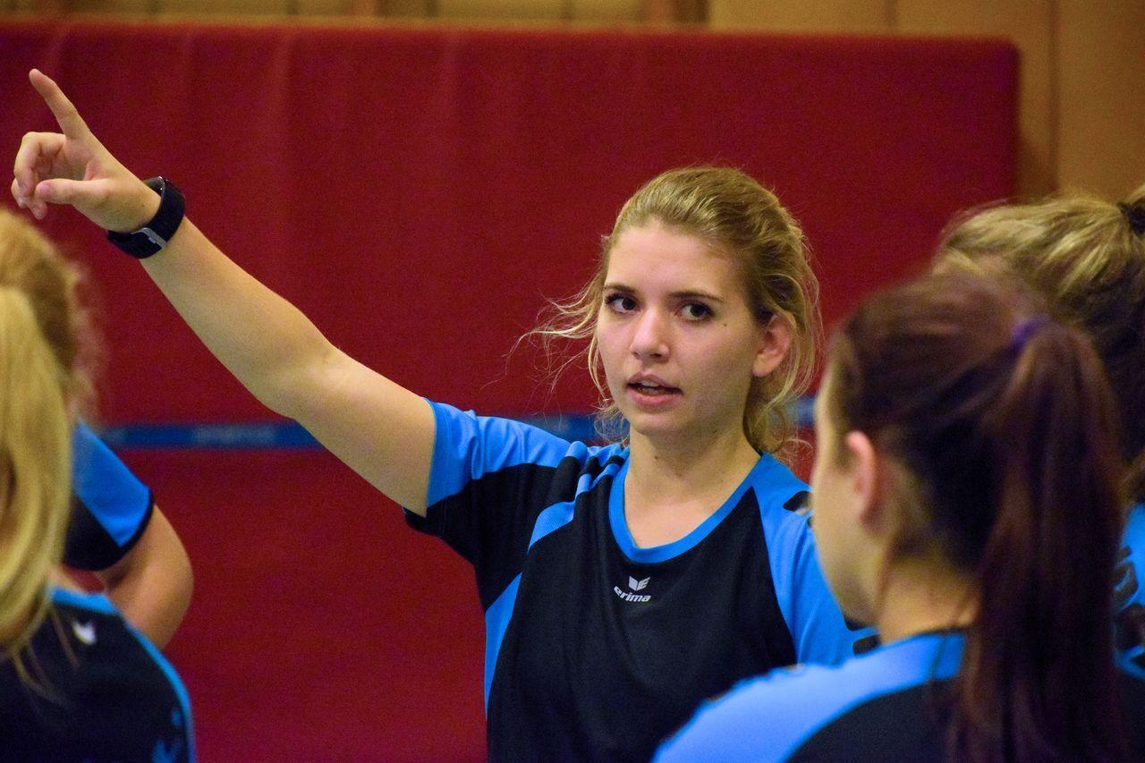 Sportlerin mit erhobener Hand erklärt etwas zu einem Team im Training, umgeben von anderen Spielerinnen in Sportkleidung.