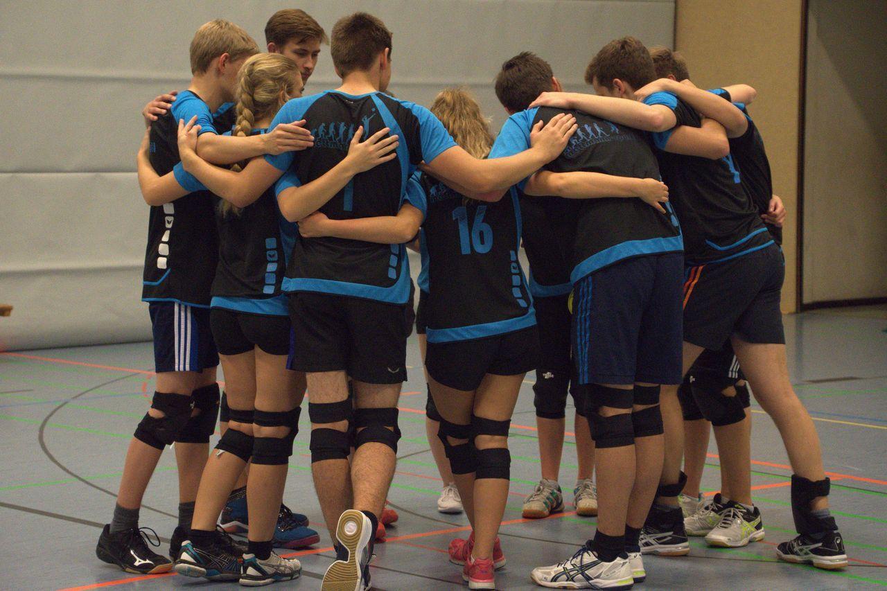 Gruppenumarmung von Jugendlichen in Sportkleidung vor einer Turnhallenwand während des Mannschaftstrainings.