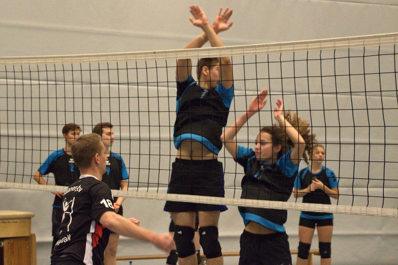 Zwei Volleyballspieler blocken am Netz, während ein dritter Spieler im Hintergrund den Ball anspielt.